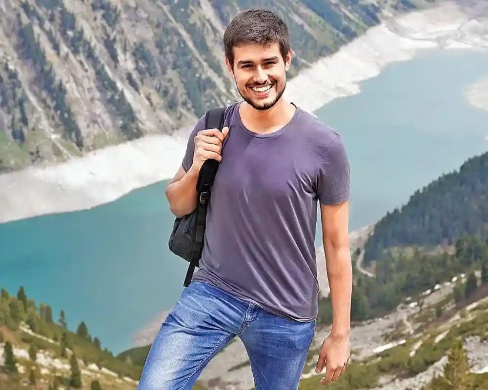Dhruv Rathee smiling with backpack at scenic mountain lake viewpoint.