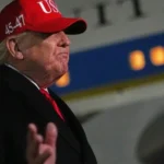 Donald Trump in a red cap speaking at an event, with his face blurred in the image.