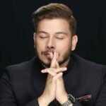Nitish Rajput posing in a dark suit with folded hands against a black background.