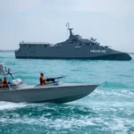 A military patrol boat in the foreground with a larger naval vessel in the sea near the Strait of Hormuz.
