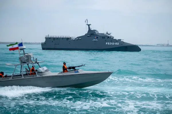 A military patrol boat in the foreground with a larger naval vessel in the sea near the Strait of Hormuz.