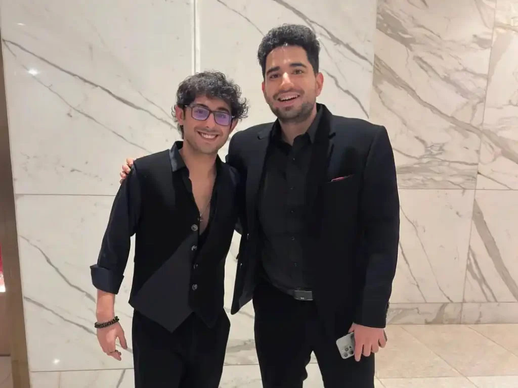 Samay Raina and CarryMinati posing together in black formal wear against a marble wall.