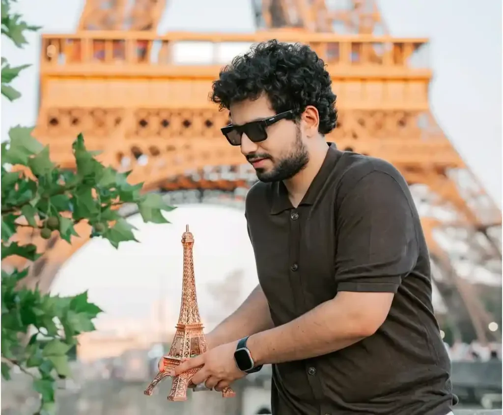 Samay Raina holding a small Eiffel Tower model in front of the real Eiffel Tower outdoors.