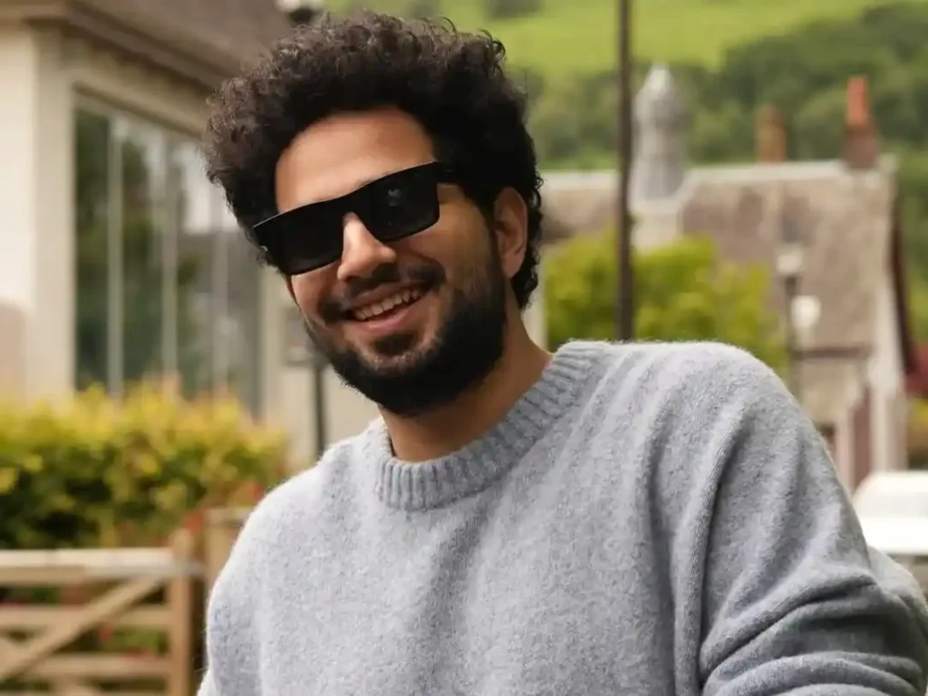 Samay Raina smiling in a grey sweater outdoors near a countryside house with green hills.