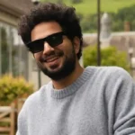 Samay Raina smiling in a grey sweater outdoors near a countryside house with green hills.