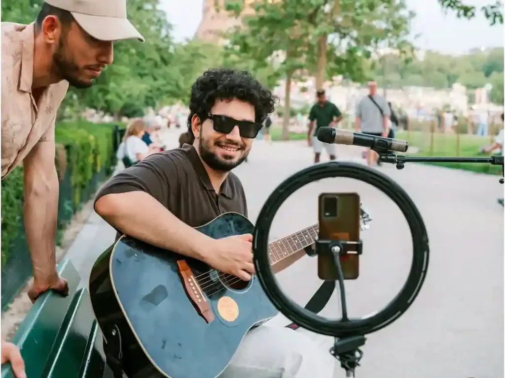 Samay Raina playing guitar outdoors during a recorded shoot with a phone and ring light in front of him.