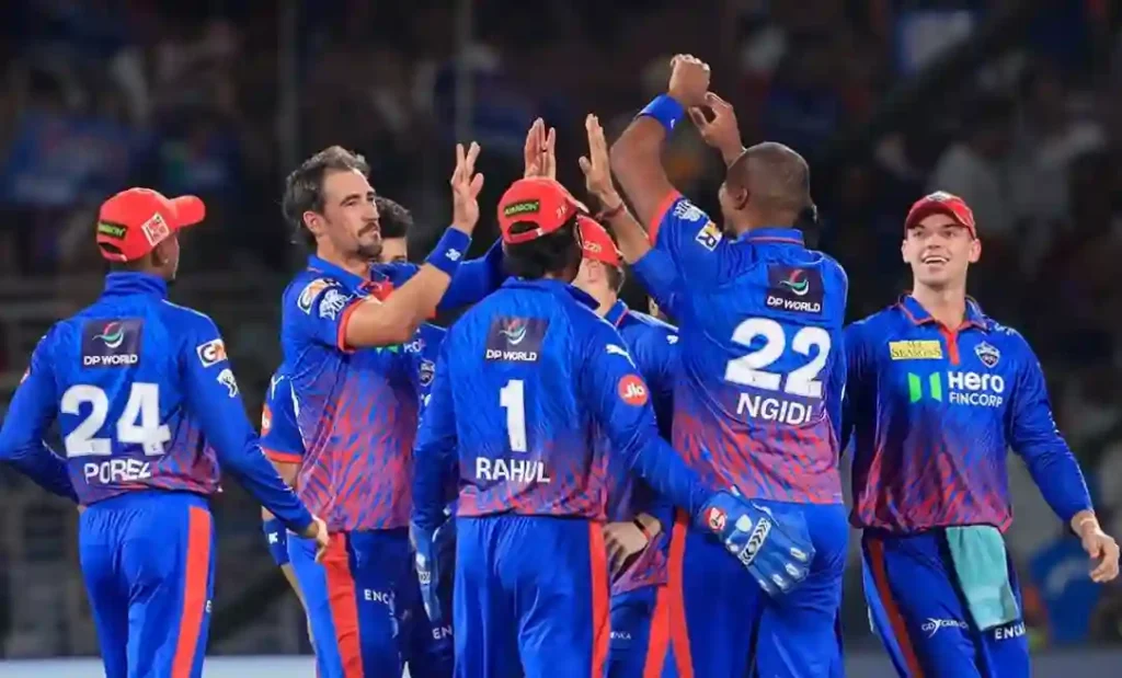 Delhi Capitals players in their blue and red jerseys, including Mitchell Starc, KL Rahul, and Lungi Ngidi, high-fiving and celebrating together on the cricket field.