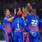 Delhi Capitals players in their blue and red jerseys, including Mitchell Starc, KL Rahul, and Lungi Ngidi, high-fiving and celebrating together on the cricket field.
