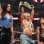Liv Morgan and Roxanne Perez holding up championship belts in a WWE Raw ring alongside Raquel Rodriguez.
