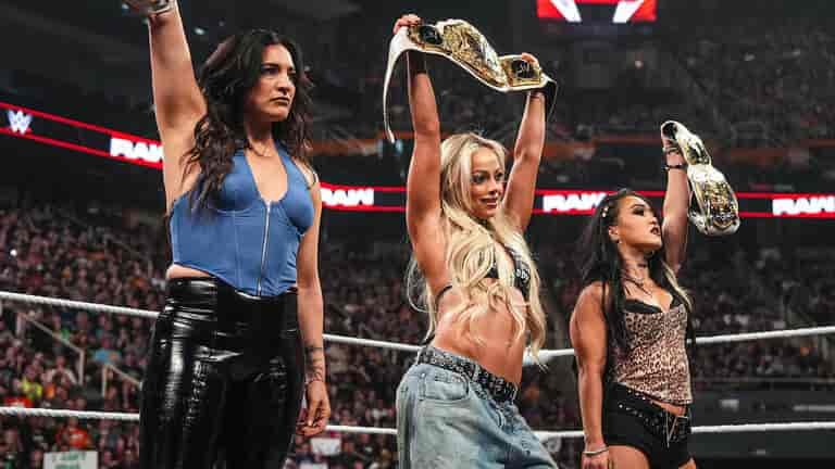 Liv Morgan and Roxanne Perez holding up championship belts in a WWE Raw ring alongside Raquel Rodriguez.